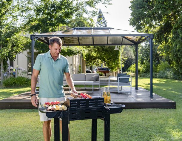A man-making barbecue in front of a Martinique 10'x14' aluminum grey gazebo with polycarbonate roof panels on a deck patio with garden furniture