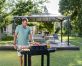 A man-making barbecue in front of a Martinique 10'x14' aluminum grey gazebo with polycarbonate roof panels on a deck patio with garden furniture